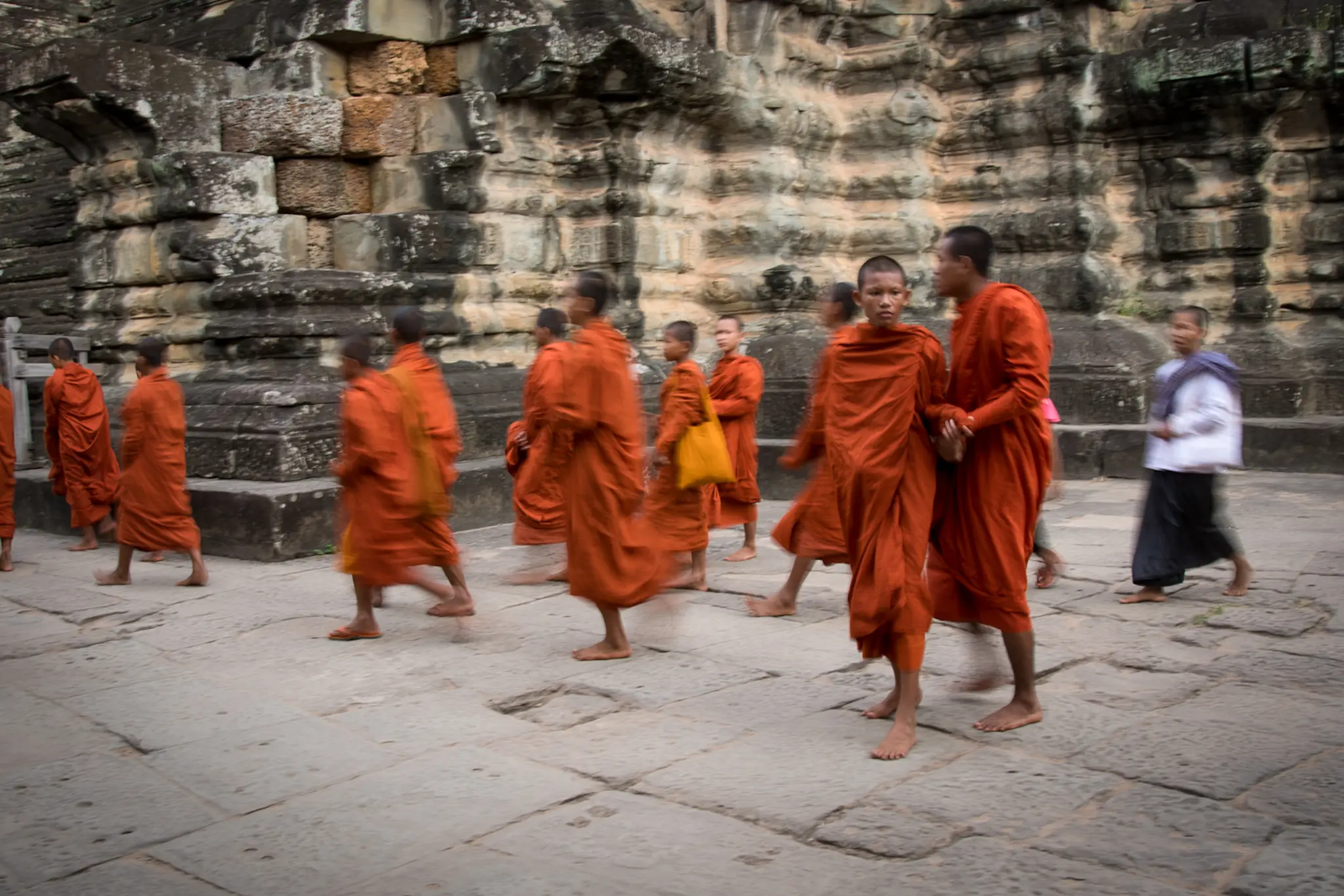 A line of young monks in orange robes walking across the stones at Angkor
